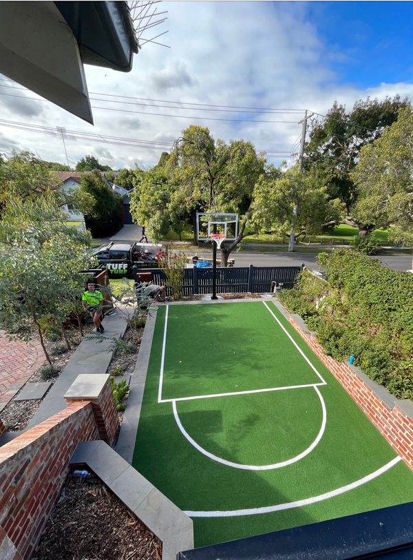 Synthetic turf backyard basketball court with hoop, surrounded by brick and greenery