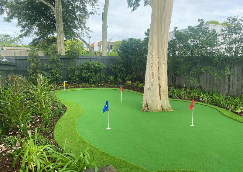 Backyard artificial putting green with golf flags, surrounded by plants and trees