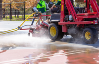 A red SMG WashMatic machine with a mounted scrubber is operating outdoors on a wet paved surface, with operators visible in the cab and yellow hoses connected. The scene includes a real outdoor environment with trees and fencing in the background.