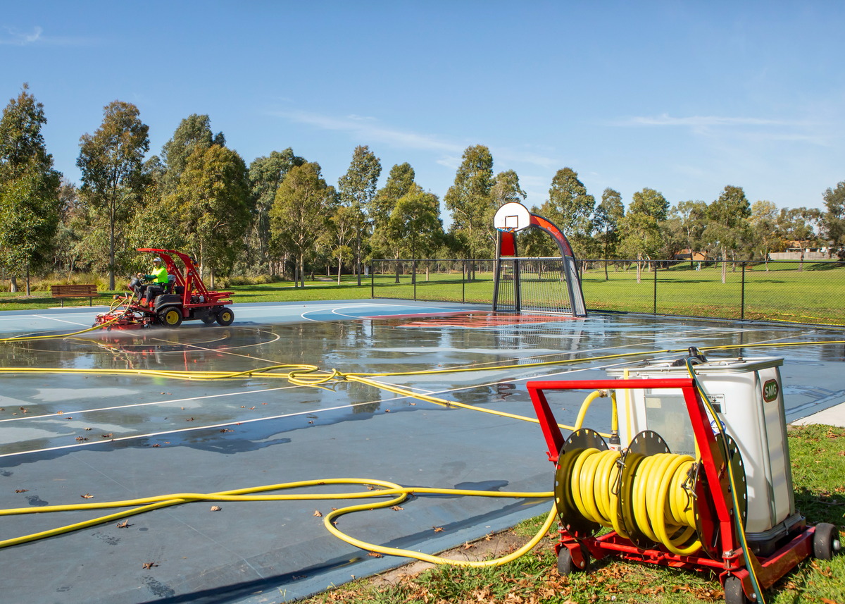 Outdoor basketball court being cleaned with a yellow hose connected to a red wheeled water tank, with a red SMG WashMatic machine and trees in the background.