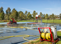 Outdoor basketball court being cleaned with a yellow hose connected to a red wheeled water tank, with a red SMG WashMatic machine and trees in the background.